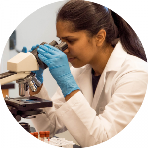 brown woman wearing a labcoat and looking through a microscope